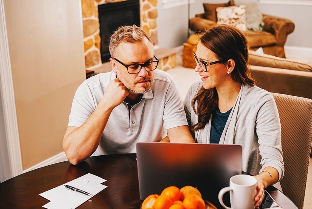 Couple discussing event planning over a laptop at home, with notes and a bowl of oranges on the table, illustrating budget preparation and organization for successful event management.