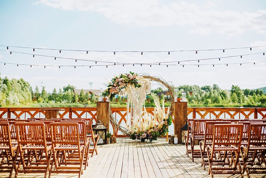 Wedding ceremony setup at a riverbank venue featuring wooden chairs, floral arch, and decorative string lights, emphasizing event planning and celebration themes.