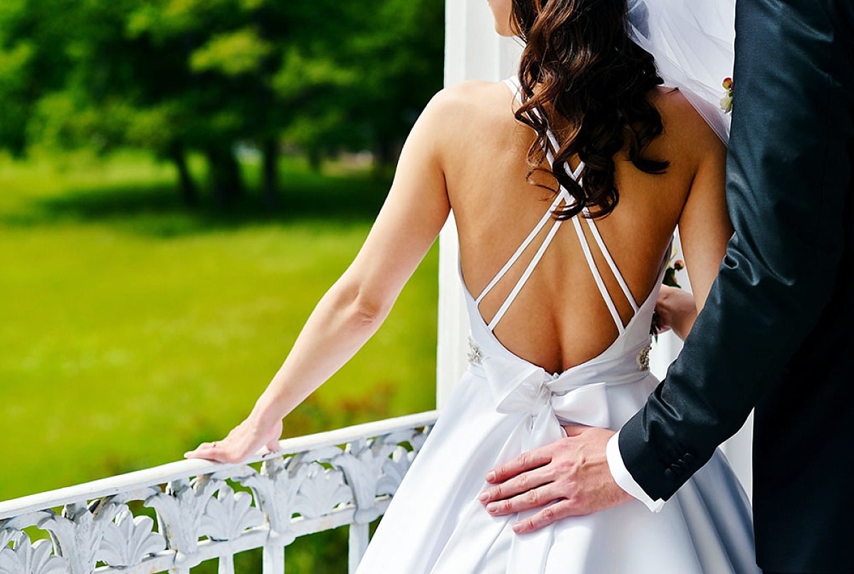 Wedding couple embracing outdoors, bride in elegant white dress with lace veil, groom in black suit, romantic setting with greenery in background.