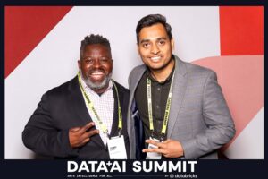 Two men posing together at the Data + AI Summit, smiling and making peace signs, with a branded backdrop featuring the event name and logo.