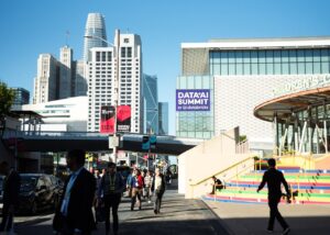 Crowd of attendees walking towards the Data + AI Summit venue with modern buildings in the background, showcasing the event's branding and vibrant atmosphere.