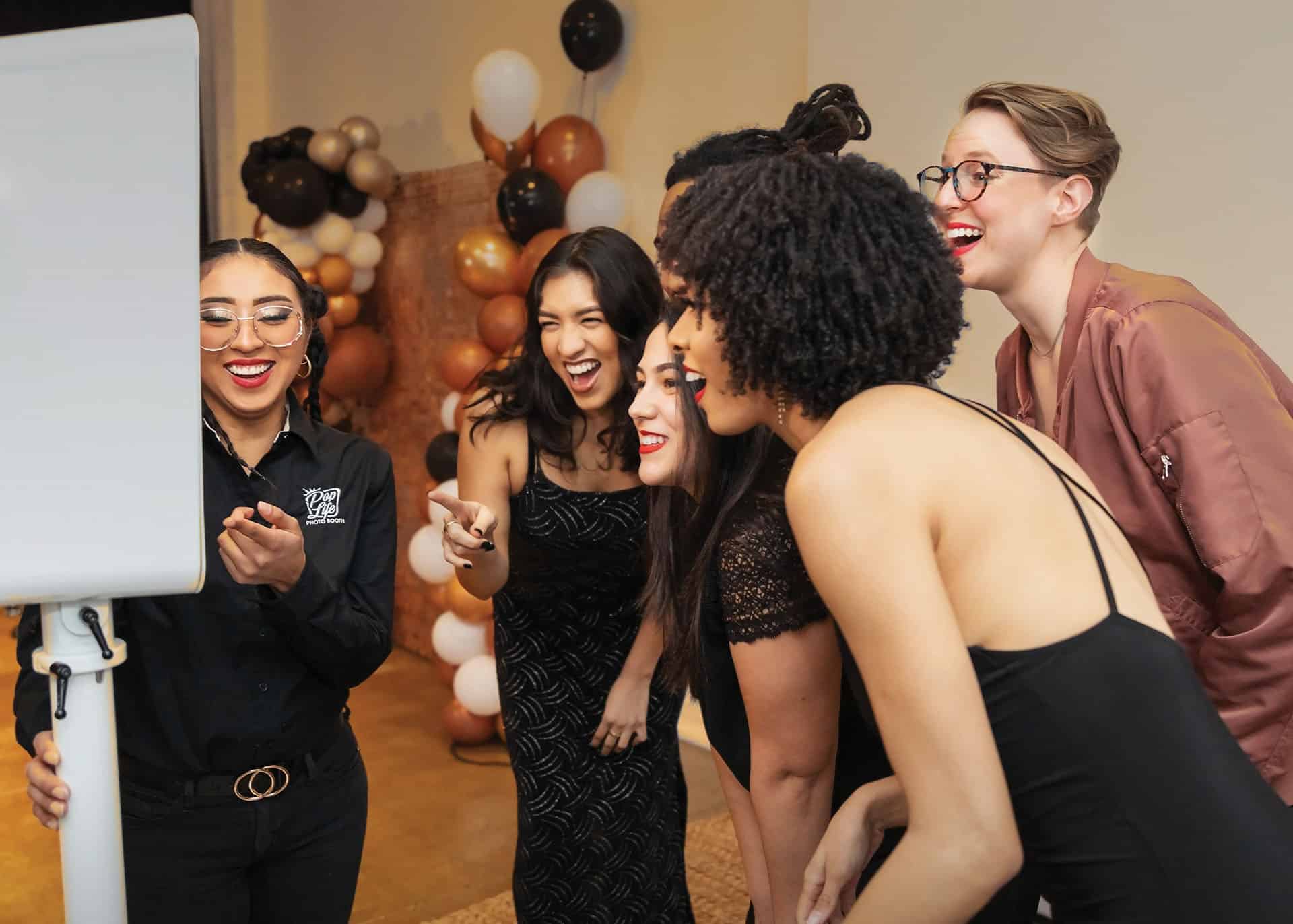 Group of women laughing and interacting joyfully around a photo booth, with a Pop Life Photo logo visible, showcasing a fun event atmosphere enhanced by the photo booth experience.