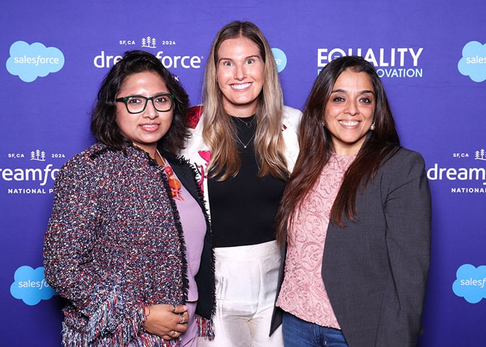 Three women posing together in front of a Salesforce Dreamforce backdrop, showcasing a vibrant atmosphere of the event, emphasizing engagement and celebration.