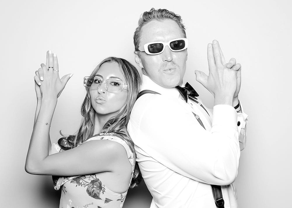Couple posing in a playful Charlie's Angels stance, making finger guns and wearing stylish sunglasses, showcasing a fun photo booth moment.