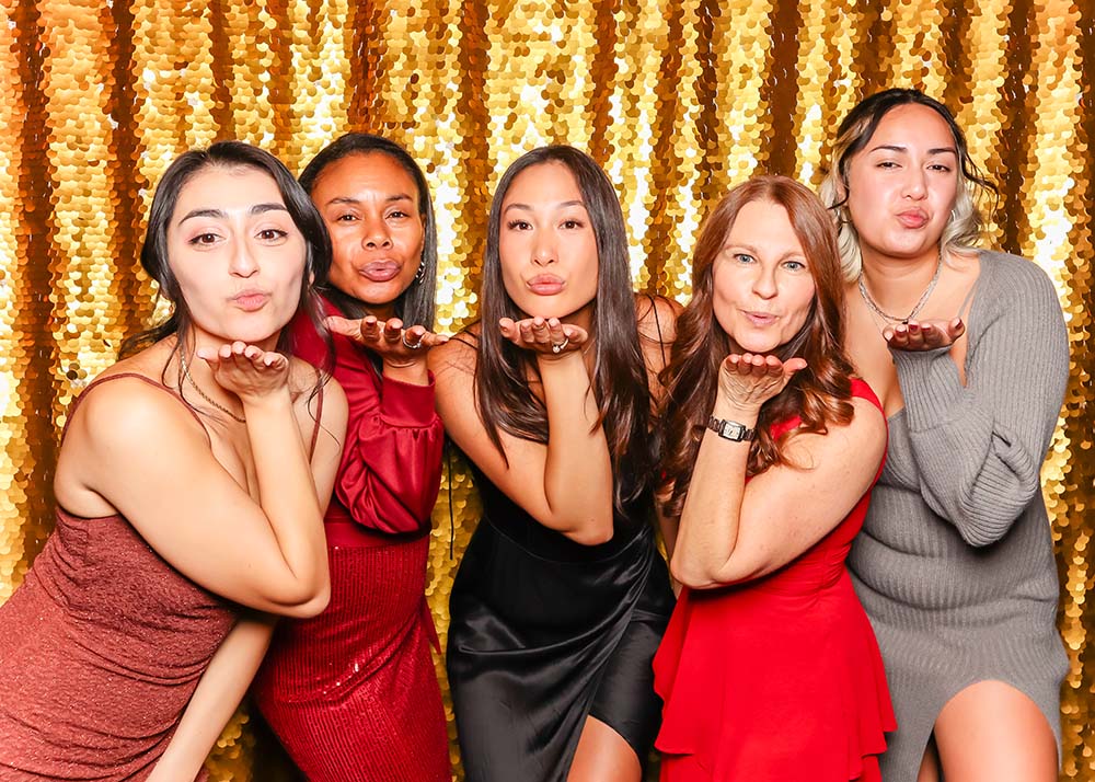 Group of five women blowing kisses in a photo booth, posing playfully against a shimmering gold backdrop, capturing the popular "blow a kiss" pose for memorable event photography.