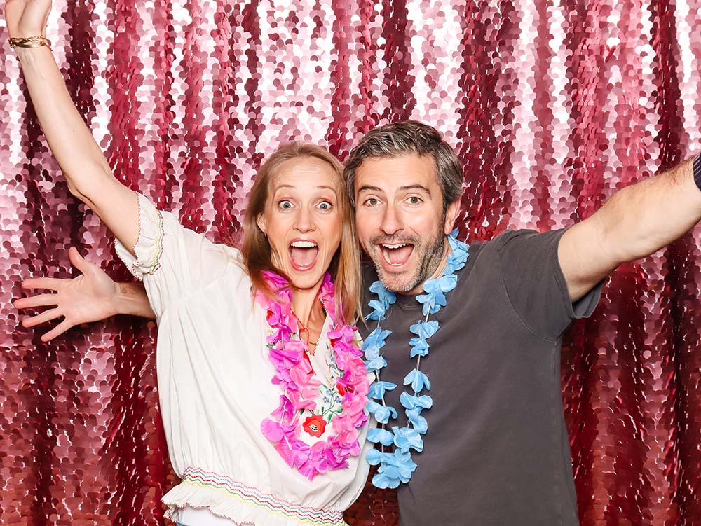 Happy couple posing in front of a sparkling pink sequin backdrop, wearing floral leis, celebrating a fun event, ideal for personalized photo opportunities and brand integration.