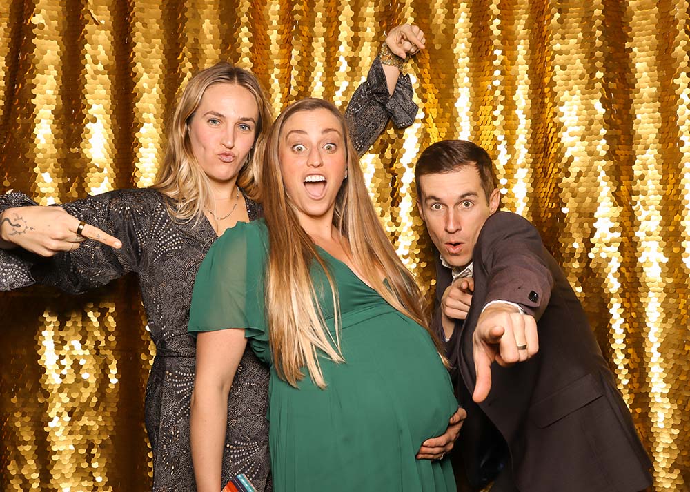 Group of three friends posing playfully in front of a shimmering gold backdrop, with one person pointing at the camera, another giving a playful gesture, and the pregnant woman in the center smiling joyfully, reflecting fun photo booth moments.