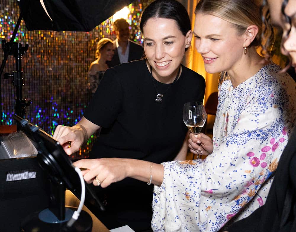 Two women interacting with a photo booth touchscreen, one holding a glass of champagne, in a vibrant event setting with colorful background lights.