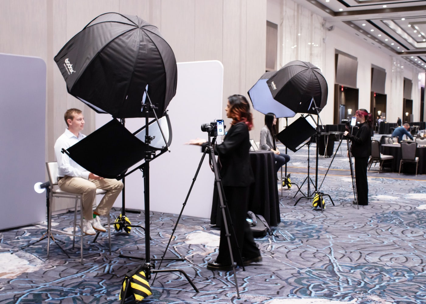 Professional headshot photography setup with lighting equipment, photographer directing a subject, and multiple headshot stations in a corporate event setting.