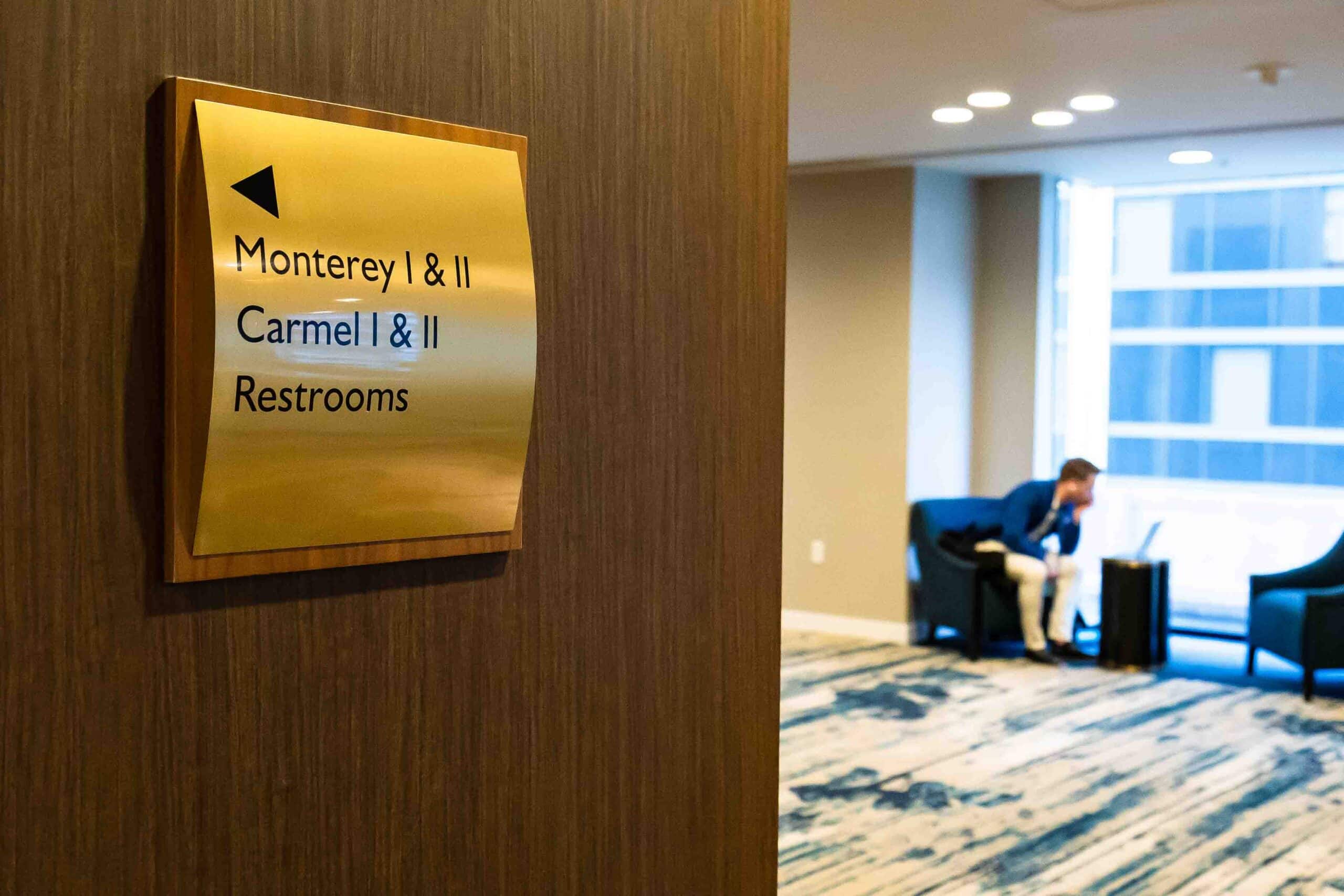 Signage indicating restrooms and meeting rooms at Hotel Nikko San Francisco, with a blurred figure seated nearby in a modern lounge area.