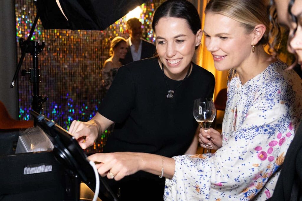 Two women interacting with a photo booth touchscreen at a vibrant event, with a sparkling backdrop and attendees in the background, highlighting the engaging atmosphere of Hotel Nikko San Francisco's event services.
