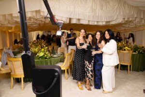 Group of four women posing with drinks in a festive event setting, featuring a photo booth in the foreground, emphasizing social interaction and engagement at the event.