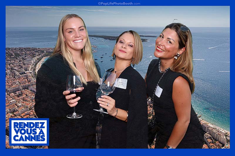Three women in elegant black outfits holding glasses, posing in front of a scenic coastal backdrop with the text "RENDEZ VOUS À CANNES," representing a fun and engaging photo booth experience at an event.