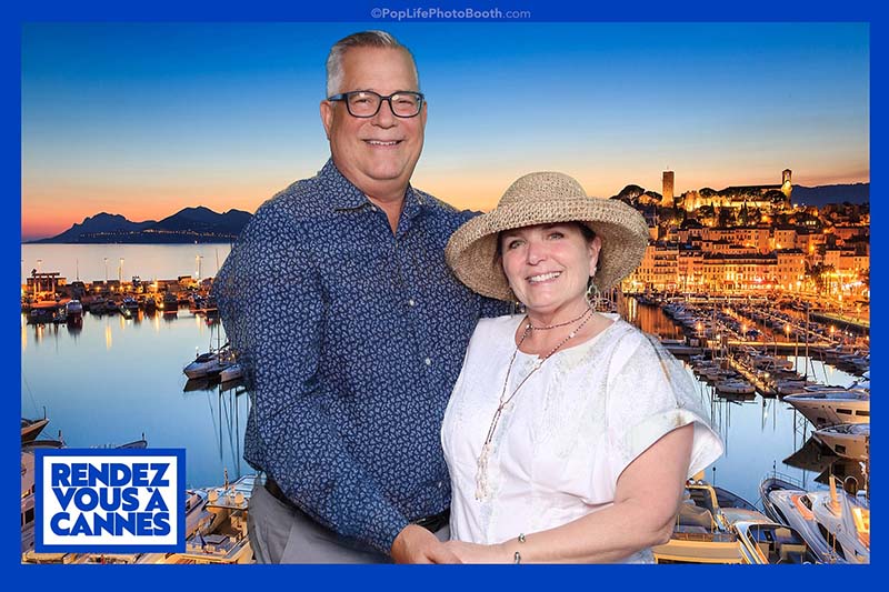Couple posing in a photo booth with a scenic waterfront background, wearing summer attire and a straw hat, featuring a “RENDEZ VOUS À CANNES” logo, capturing joyful moments at a wedding event.