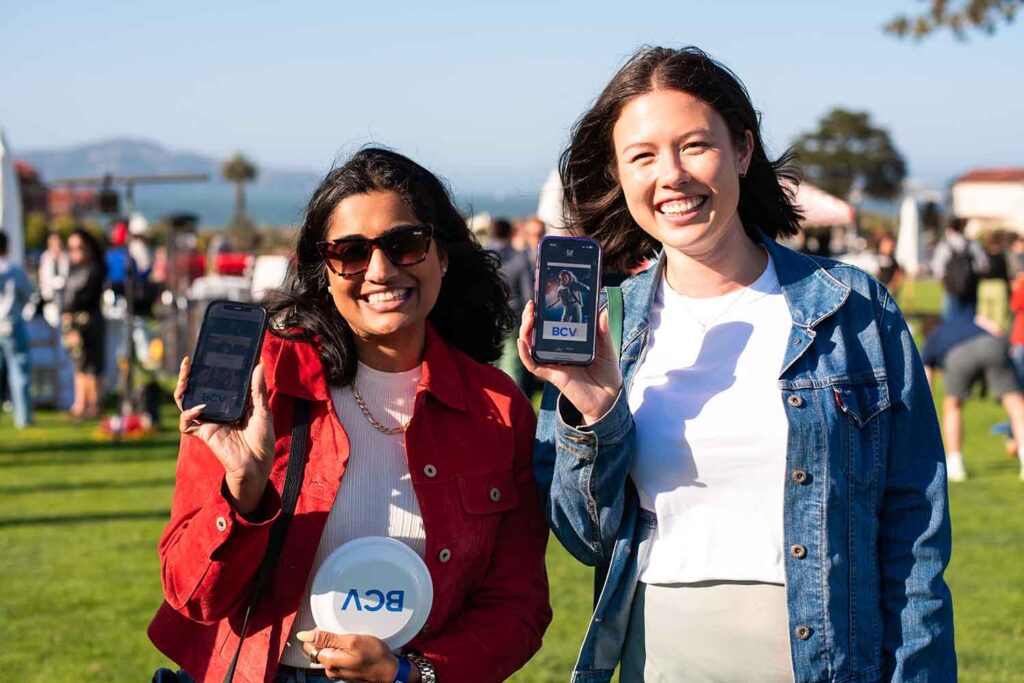 Two women smiling and holding smartphones displaying photos, with a summer event setting featuring grass and distant palm trees, capturing the fun of a summer wedding photo booth experience.