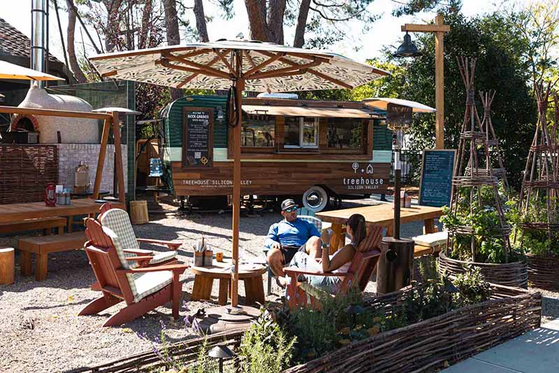 Couple enjoying drinks under a patterned umbrella at Treehouse Hotel Silicon Valley, with a rustic food truck and charming outdoor seating, surrounded by greenery and whimsical decor.
