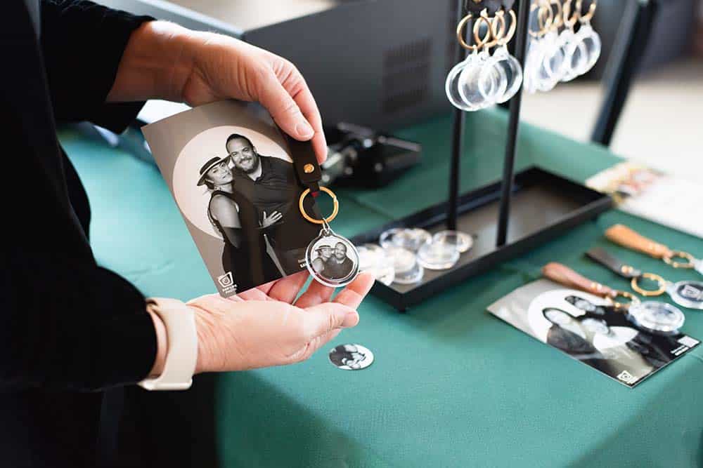 Hand holding a keychain photo souvenir featuring a couple, with a printed photo and various keychain options displayed on a green table, showcasing personalized wedding memorabilia from the Spotlight Photo Booth at the Treehouse Hotel wedding showcase.