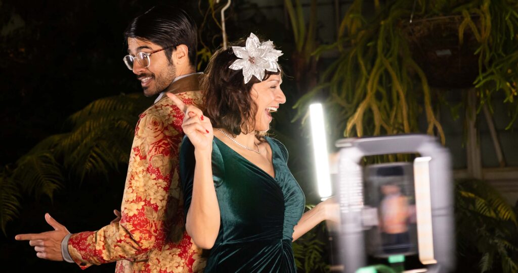 Couple posing playfully back-to-back in a vibrant event setting, showcasing retro and upscale aesthetics, with professional lighting and a camera capturing the moment.