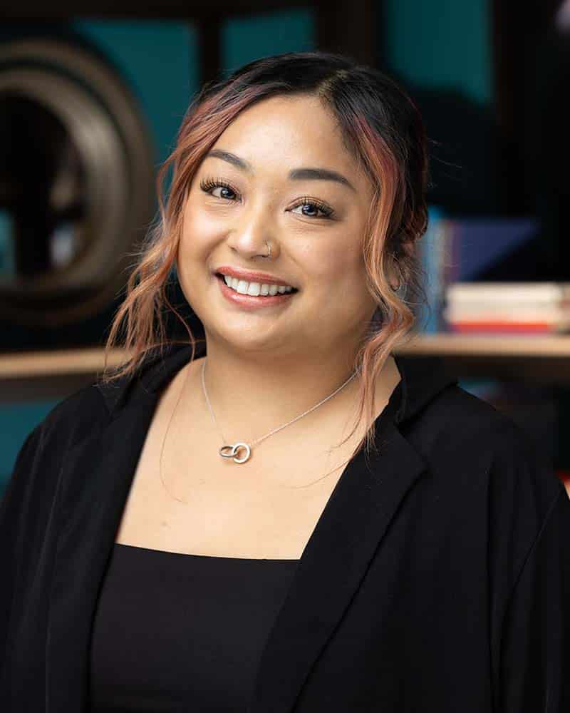 Professional headshot of a woman with a warm smile, wearing a black blazer and a silver necklace, set against a colorful background, showcasing confidence and approachability for corporate branding.