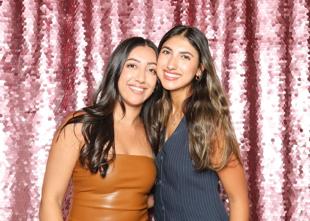 Two women smiling together in front of a shimmering pink sequin backdrop, capturing a glamorous photo booth setting ideal for celebrations and upscale events.