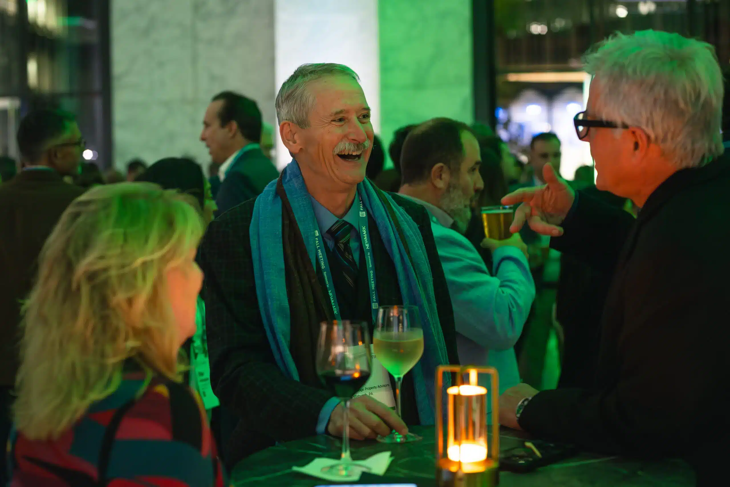 Group of people engaged in conversation at a gala event, featuring a man in a blue scarf laughing, holding a glass of wine, with green lighting creating an elegant atmosphere.