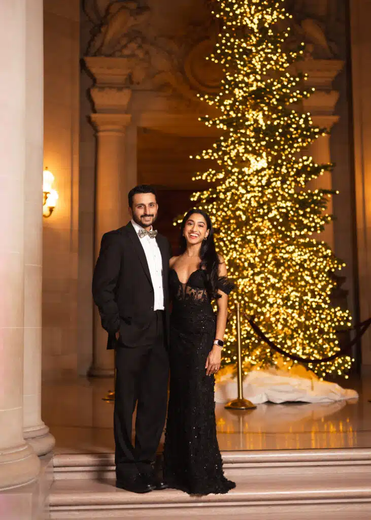 Couple posing for portrait photography at a holiday celebration with a decorated Christmas tree and elegant architecture.