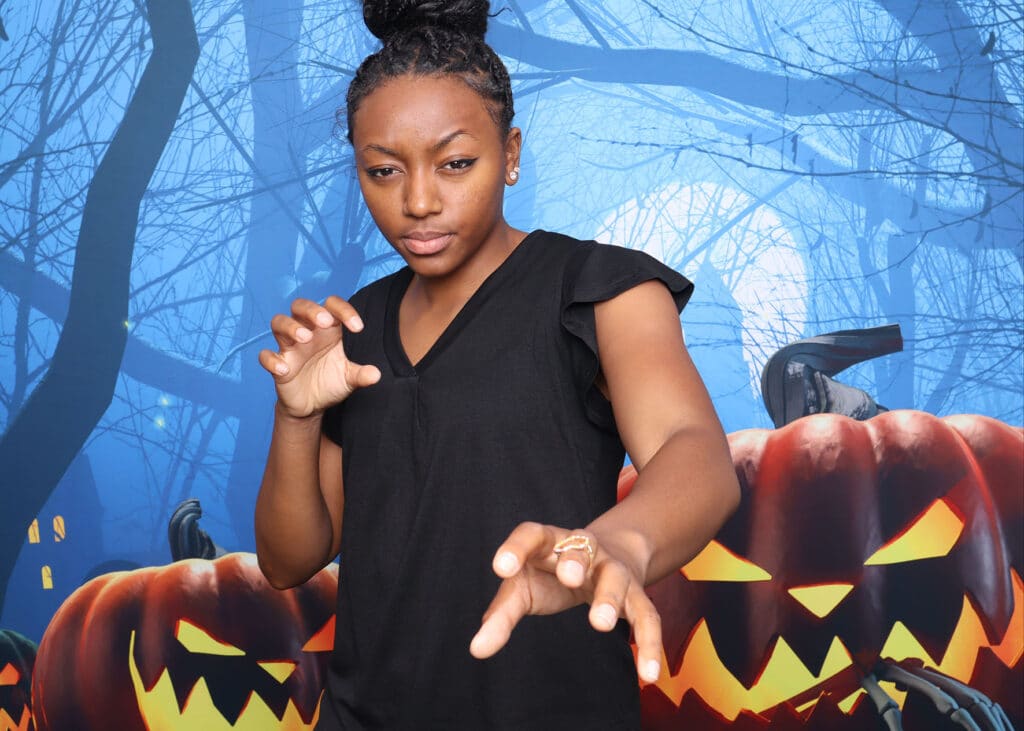 Person posing playfully in front of a spooky Halloween backdrop featuring carved pumpkins and a dark forest scene, enhancing festive photo booth experience.