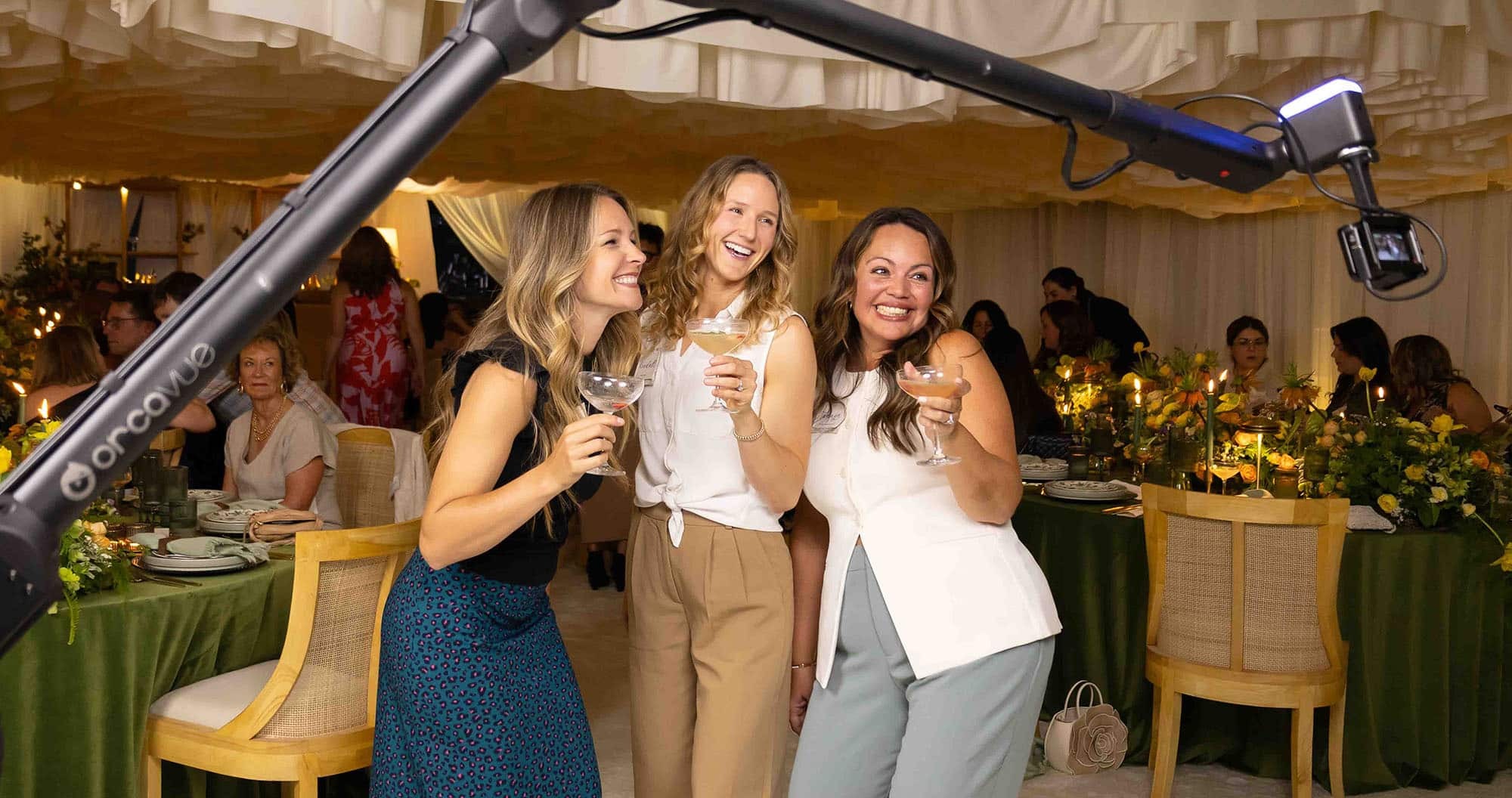 Three women joyfully posing with cocktails in front of a Glambot video booth at an elegant event, surrounded by a beautifully decorated table with floral arrangements and soft lighting.