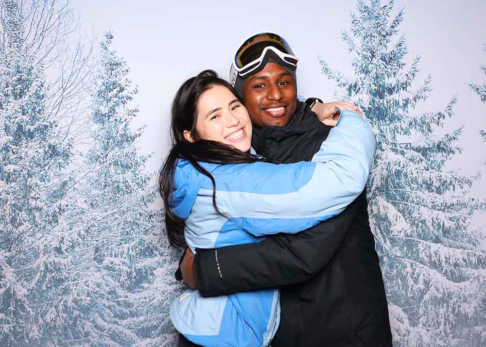 Couple embracing in winter jackets against a snowy forest backdrop, capturing a festive winter photo booth moment.