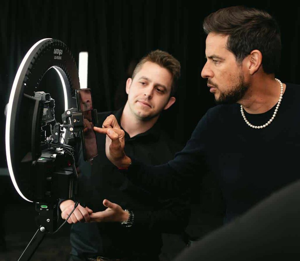 Two men interacting with a 360 video booth setup, adjusting a smartphone attached to a ring light, in a dark, elegant environment.