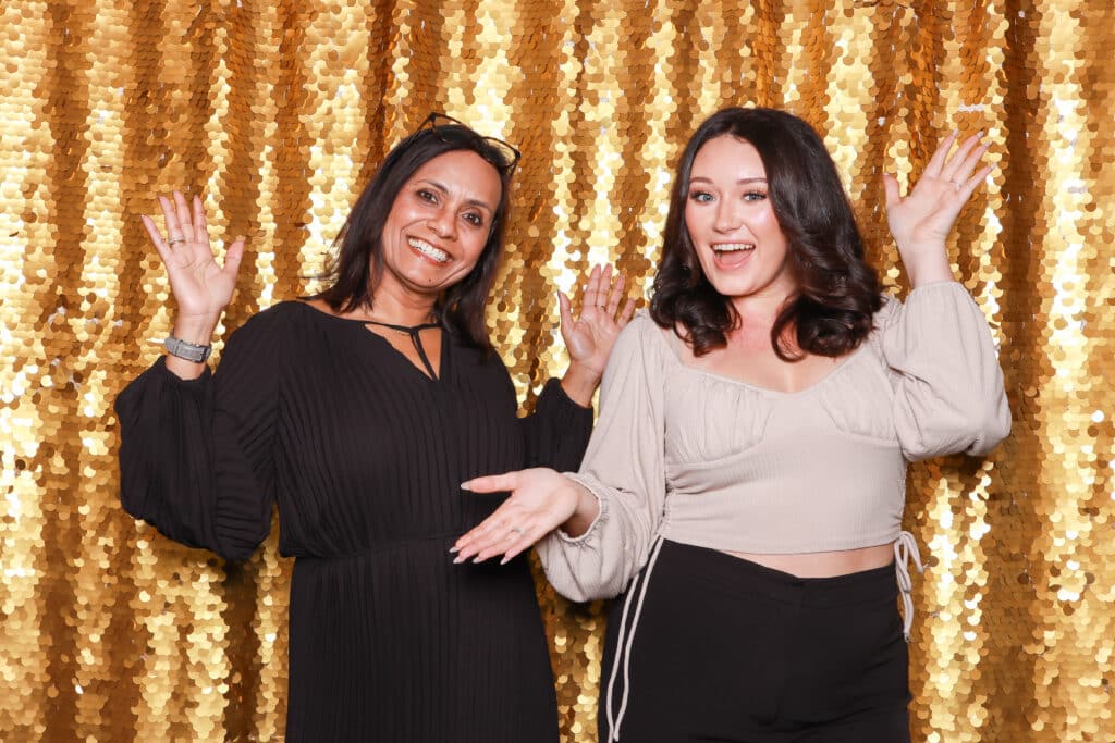 Two women posing joyfully in front of a shimmering gold sequin backdrop, ideal for glamorous photo settings at upscale events.