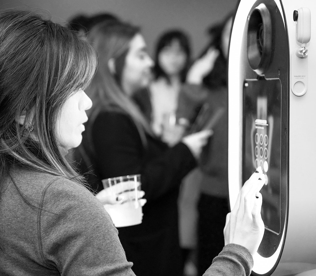 Woman interacting with a photo booth touch screen, holding a drink, surrounded by guests in a lively event setting, highlighting interactive and engaging photo experiences.
