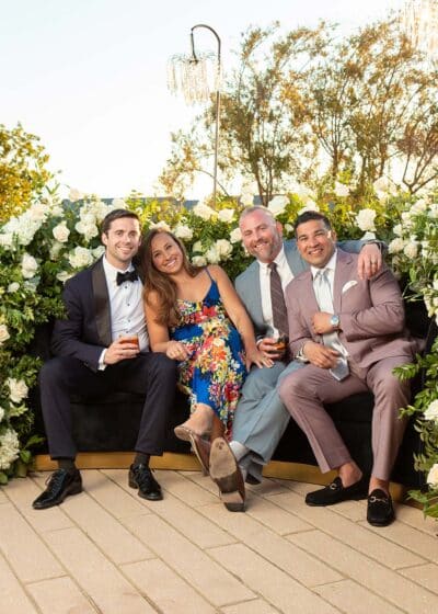 Group of four elegantly dressed individuals enjoying a luxury event, seated on a floral-adorned couch, surrounded by greenery and soft lighting, capturing a joyful moment ideal for high-end photography experiences.