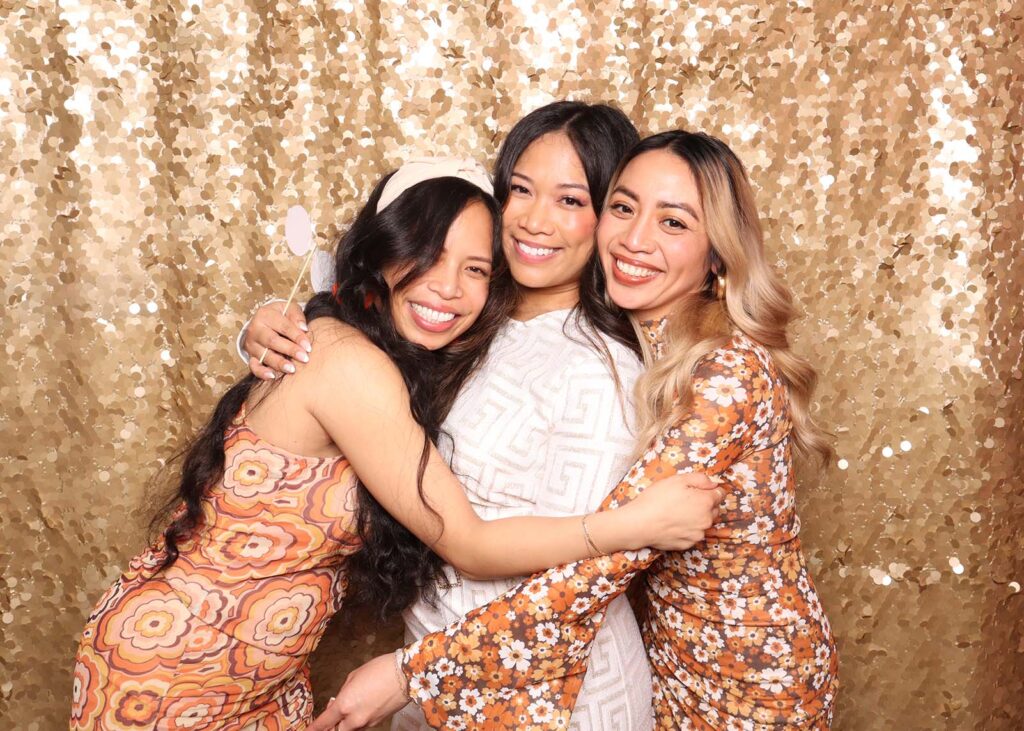 Three women embracing in front of a shimmering gold sequin backdrop, showcasing a festive and glamorous photo booth setting ideal for celebrations and upscale events.