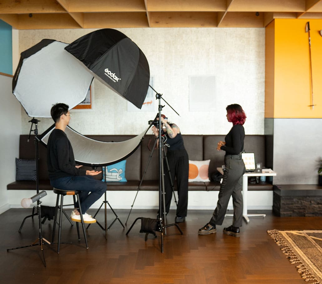 Photography session setup with a seated male model, studio lighting equipment, and two stylists adjusting the scene in a modern, well-decorated indoor space, showcasing Pop Life Photo's professional headshot service.