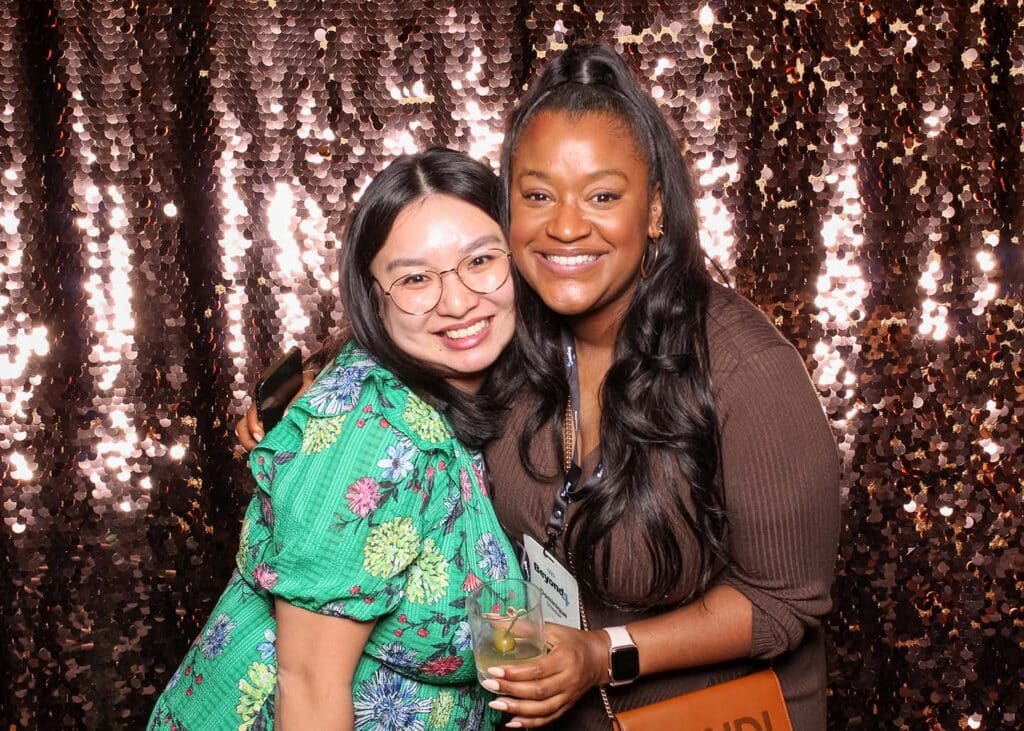 Two women smiling together in front of a shimmering sequin backdrop, reflecting light and creating a glamorous photo setting ideal for upscale events.