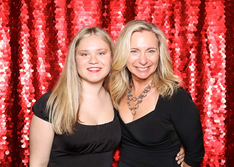 Two women smiling together in front of a vibrant red sequin photo booth backdrop, showcasing a glamorous and festive atmosphere ideal for upscale events and celebrations.