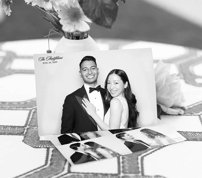 Black-and-white portrait of a couple in formal attire, smiling and posing closely, with printed photo keepsakes displayed on a table alongside a floral arrangement, capturing a glamorous event atmosphere.