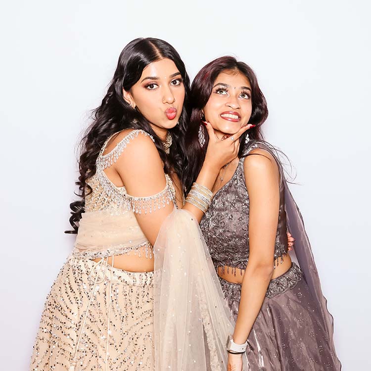 Two elegantly dressed women in glamorous attire posing playfully for a photo booth, showcasing Hollywood-inspired elegance and joyful interaction, with a white backdrop emphasizing their stylish outfits.