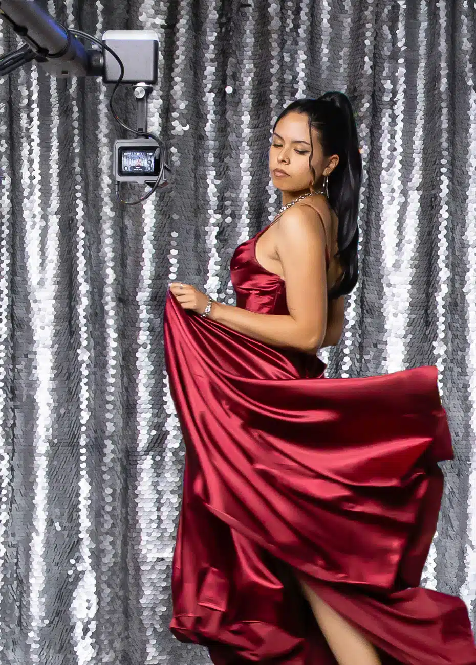 Woman in a flowing burgundy gown posing in front of a shimmering silver backdrop, showcasing the Glam Booth experience for weddings and events.