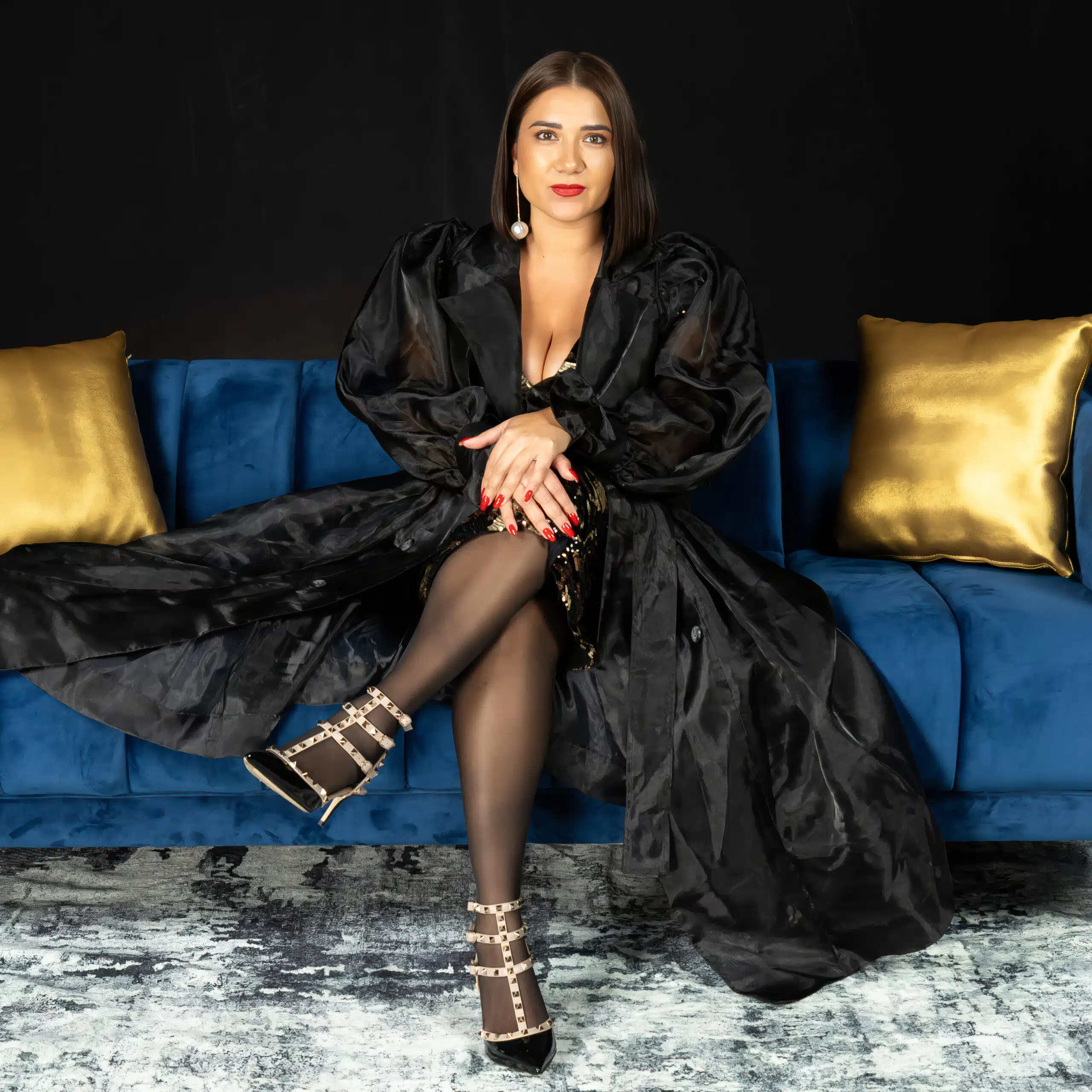 Elegant woman in a black gown seated on a blue couch, showcasing polished nails and stylish heels, embodying glamour at the Fur Ball 2025 event.
