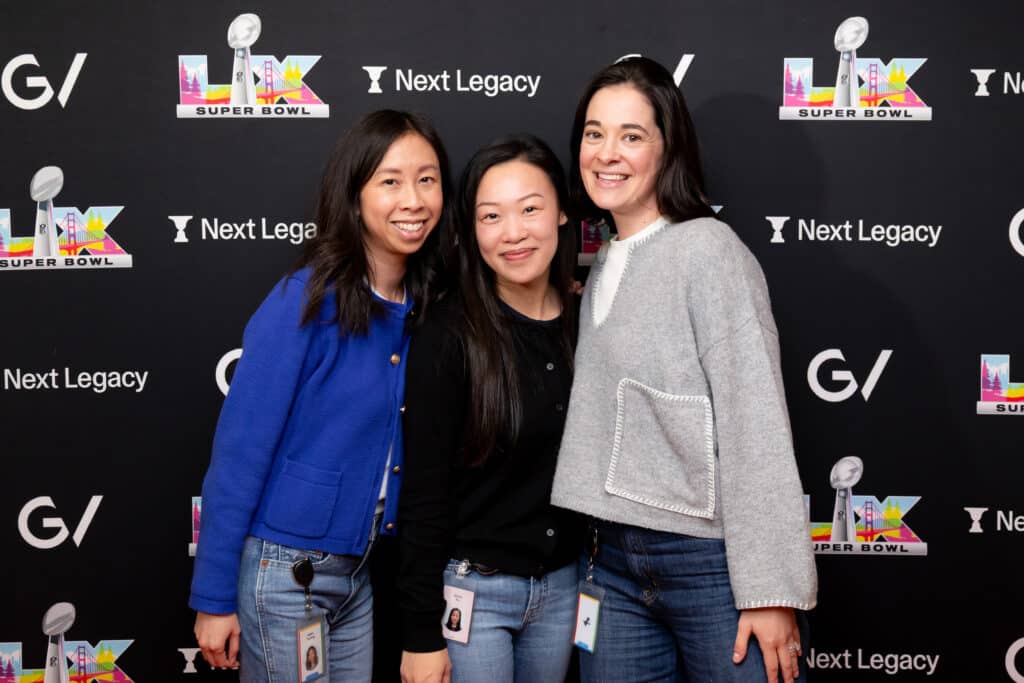 Three women posing together in front of a branded backdrop featuring the Super Bowl logo and sponsor logos, capturing a lively event atmosphere.