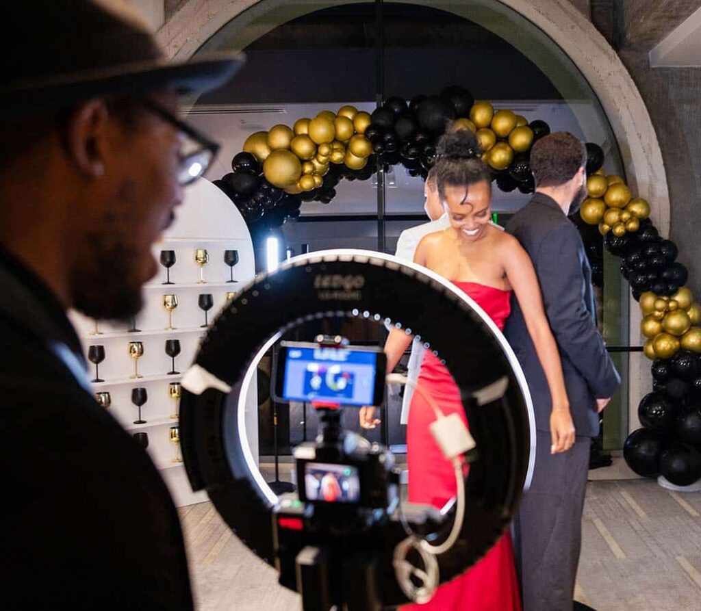 Event guests posing for a 360 Video Booth experience, featuring a woman in a red dress and a man in a suit, with a decorative balloon arch in the background.