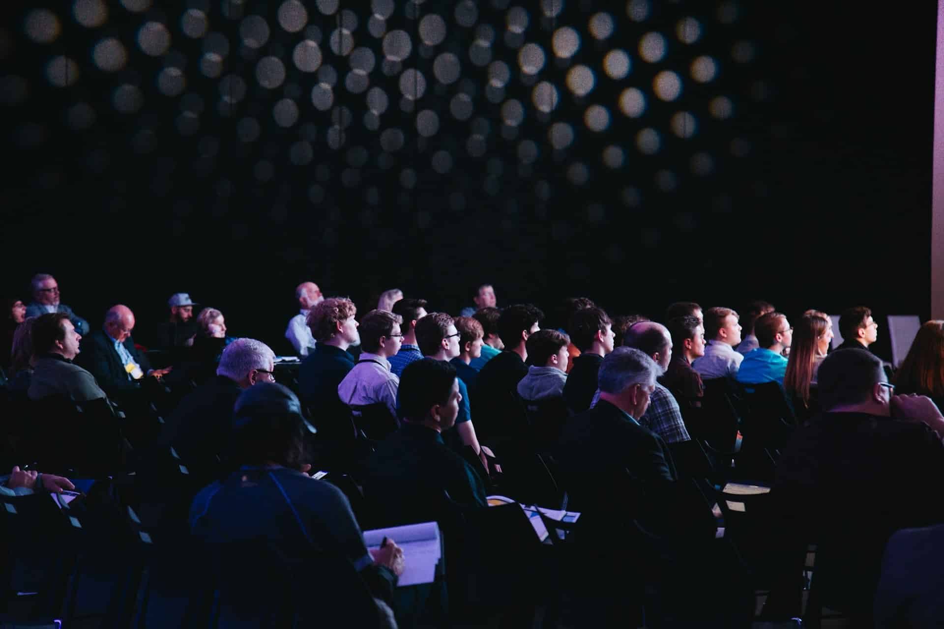 Audience engaged at a corporate event, seated in a darkened space with soft lighting and a backdrop of blurred lights, reflecting immersive trade show experiences.