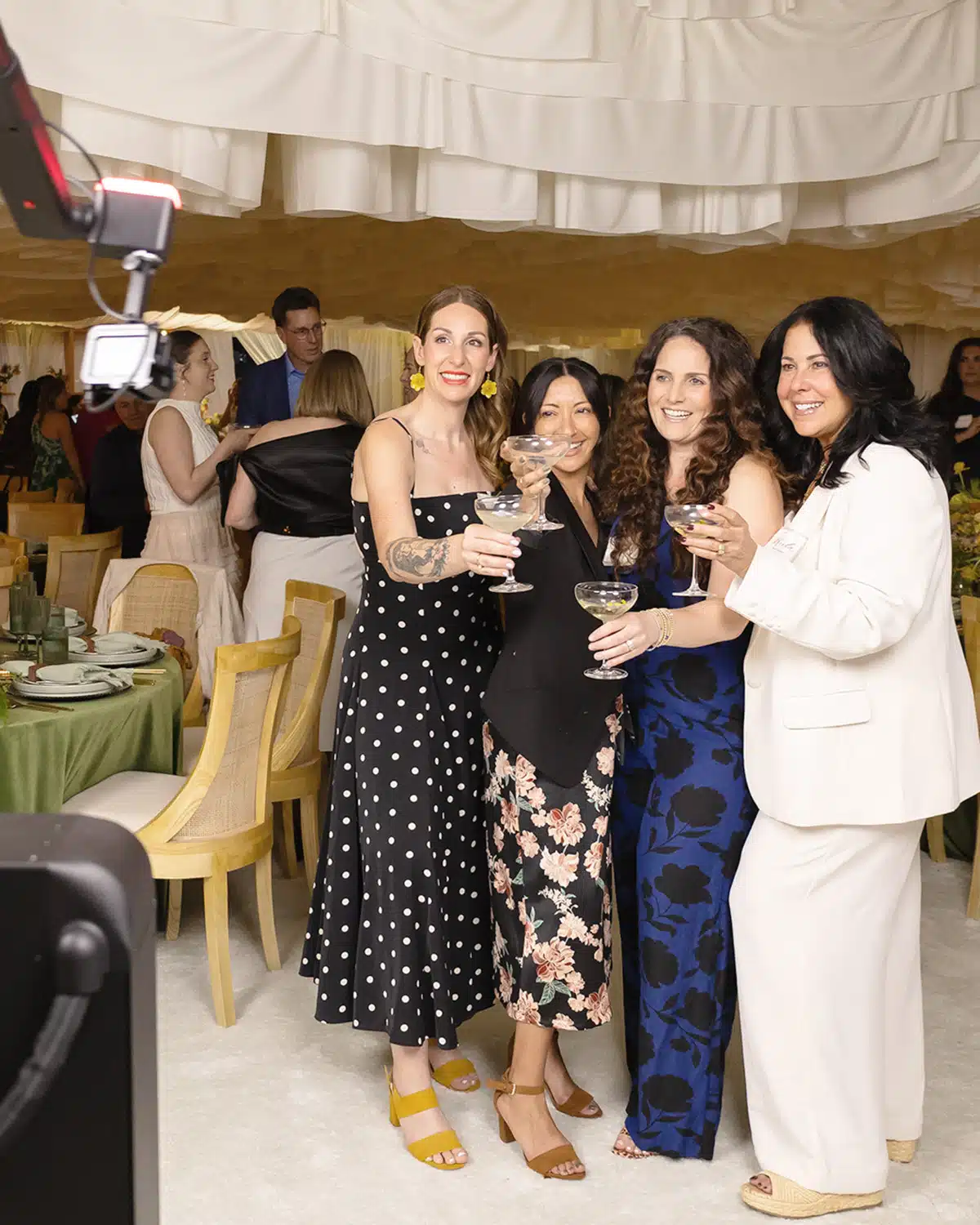 Four women cheerfully posing with cocktails in a beautifully decorated wedding venue, featuring elegant table settings and a stylish backdrop, capturing a joyful moment at a wedding celebration.