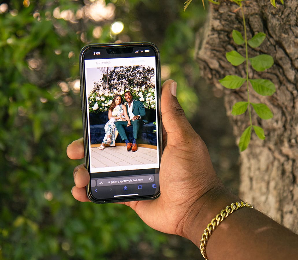 Hand holding smartphone displaying a couple posing in a professional portrait, surrounded by greenery, showcasing Pop Life Photo's photography services for events and personal portraits.