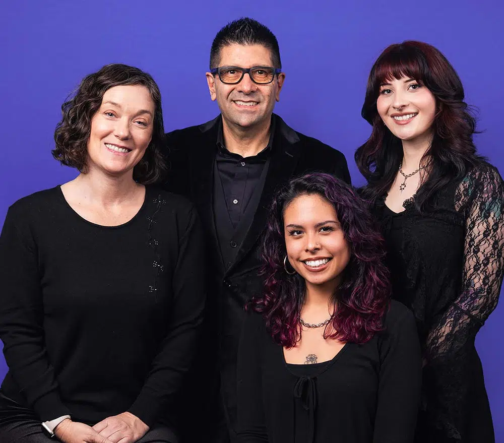 Group of four smiling individuals in black attire against a purple backdrop, representing the Pop Life Photo team, highlighting their engaging photo booth experiences.