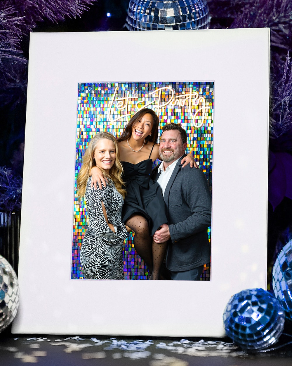 Group of three smiling guests posing in front of a colorful backdrop with "Let's Party" neon sign, showcasing a glamorous party atmosphere enhanced by disco balls and festive decor.