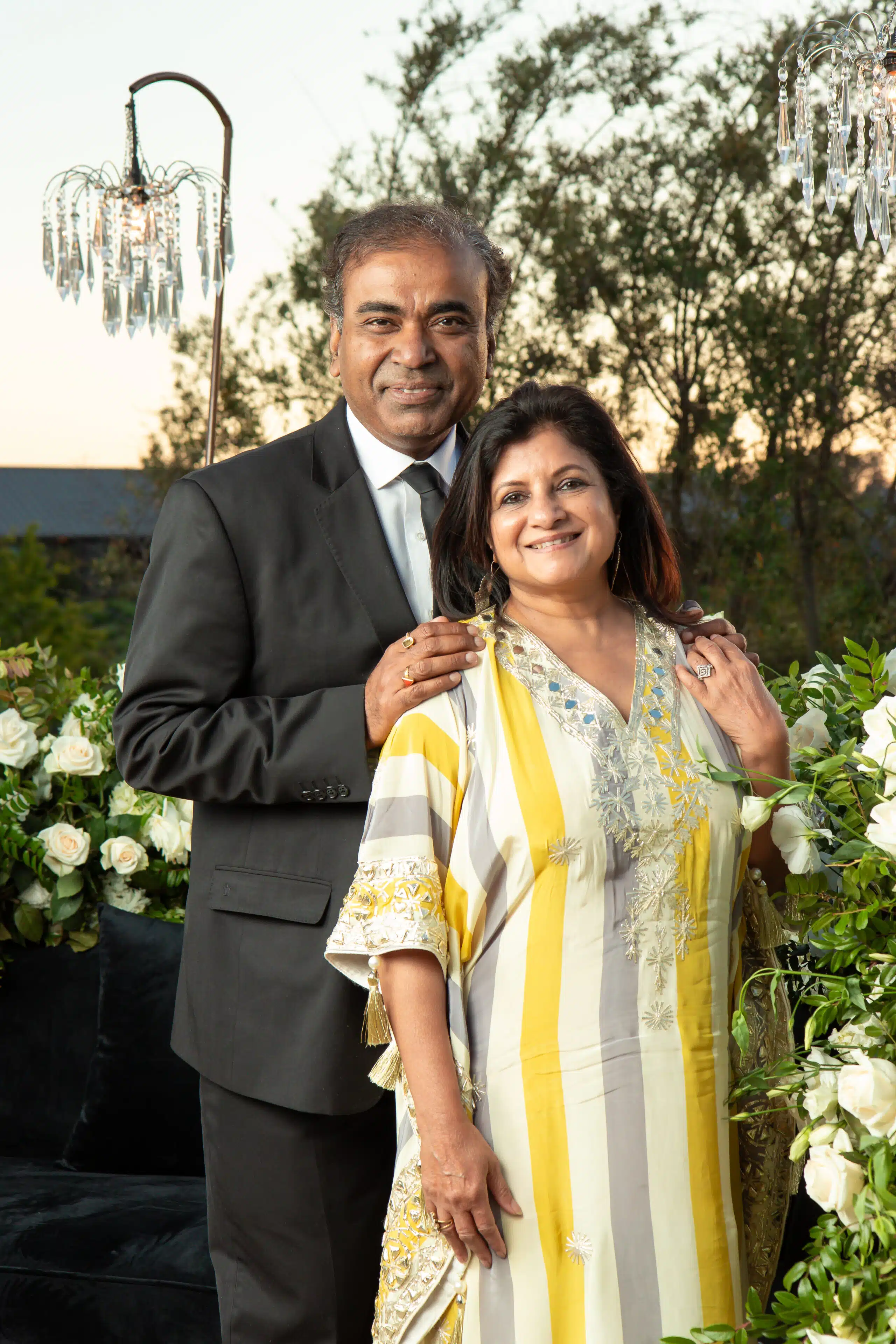 Couple posing for a portrait in formal attire, surrounded by floral arrangements and a chandelier, capturing a celebratory moment at a wedding event.