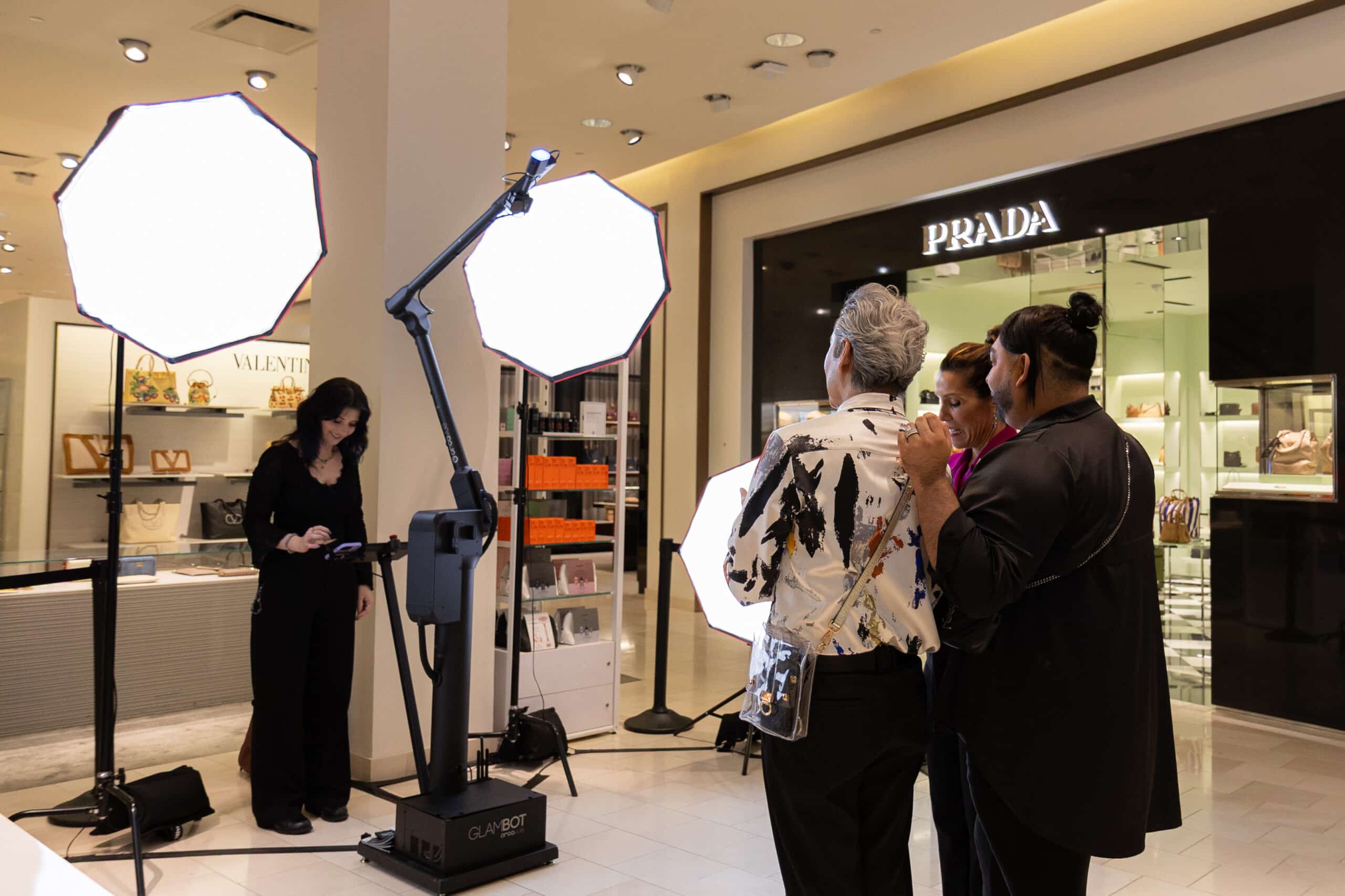 Photo booth setup with GlamBot technology, featuring guests preparing for a photo session in a stylish retail environment, illuminated by soft lighting from large octagonal lights, with luxury brand displays in the background.